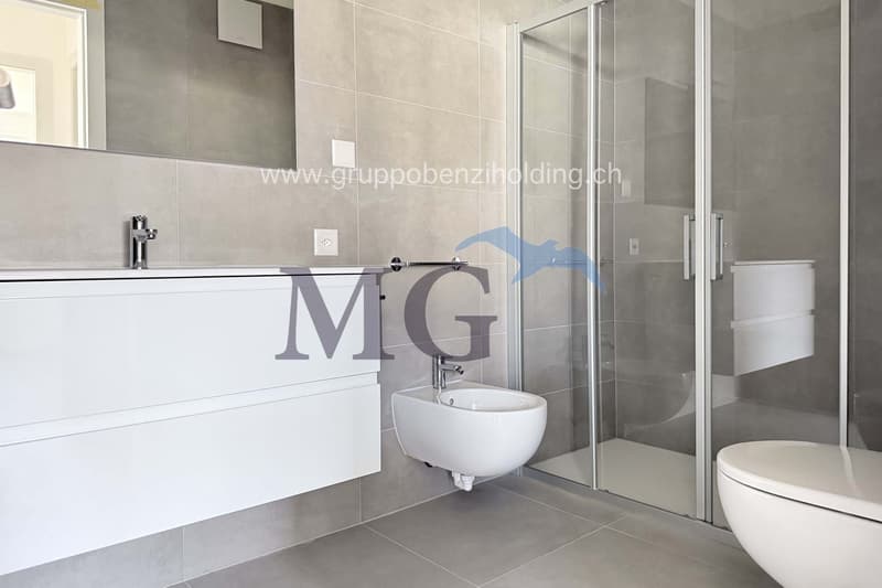 Modern bathroom with gray tiles, glass shower enclosure, white sink and vanity, bidet, toilet, faucet, and power outlet