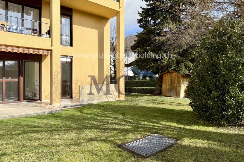 Large grass lawn, small water fountain, house entrance with balcony, hedge, small wooden storage house