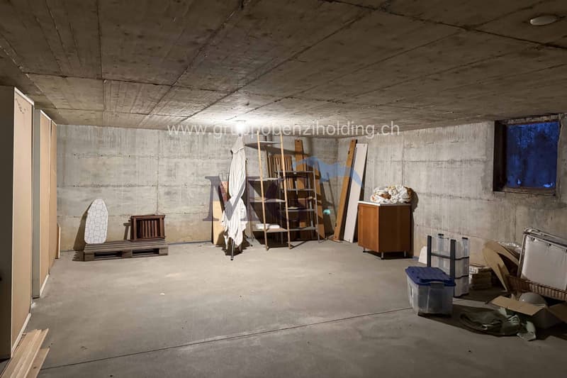 large open room with concrete walls, wooden shelves, boxes, a table, a bench and some clutter