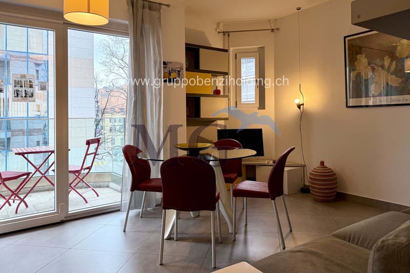 Open living area with sliding glass doors leading to balcony, wooden desk with computer, dining table with chairs, couch, side table, shelves