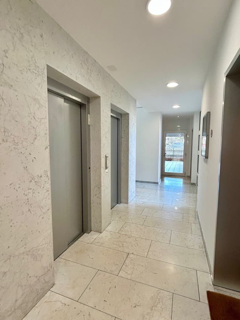 Marble walls, white tile flooring, two elevator doors, hallway leading to an open door