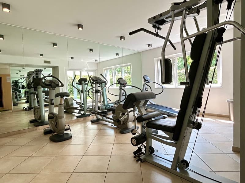 A gym room with various exercise equipment, including treadmills, stationary bikes, and weight machines. The room has large windows, ceiling lights, and a trash bin.