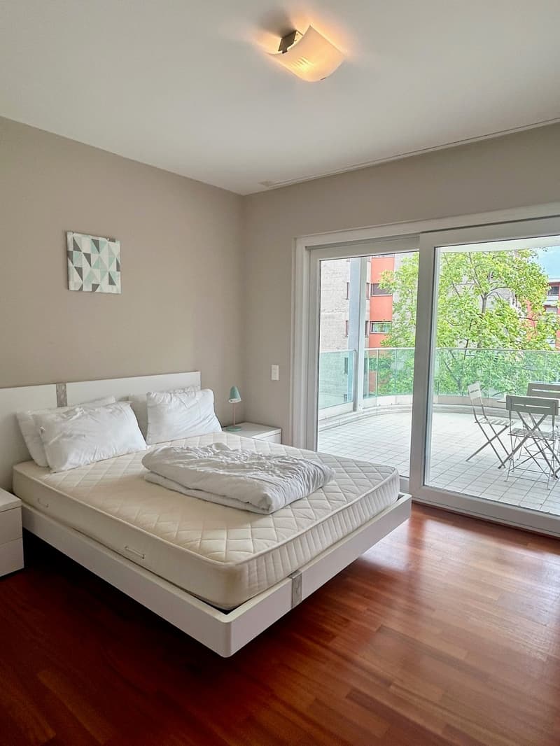 Bedroom with bed, white headboard, white bedding, nightstands, lamp, sliding glass doors, balcony with table and chairs, outdoor view
