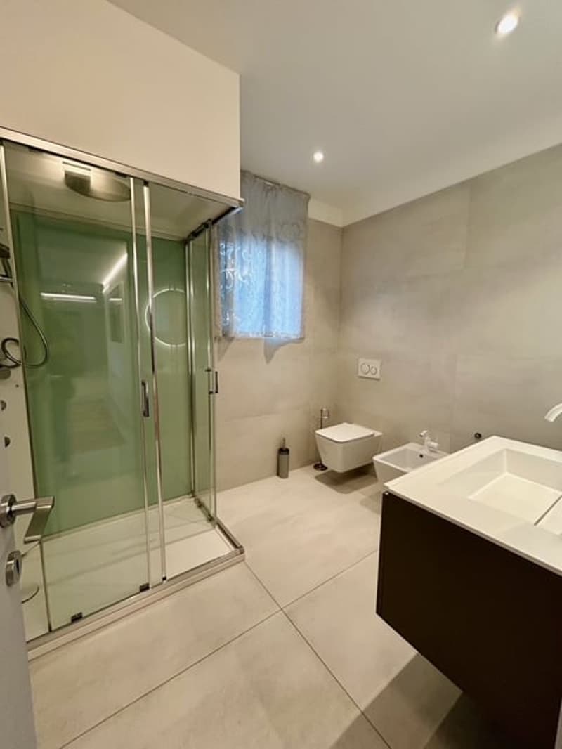 modern shower cubicle, ceramic tiled floor, white porcelain sink, dark wooden cabinet, wall mounted toilet, window with curtains, white tiles, open shower space