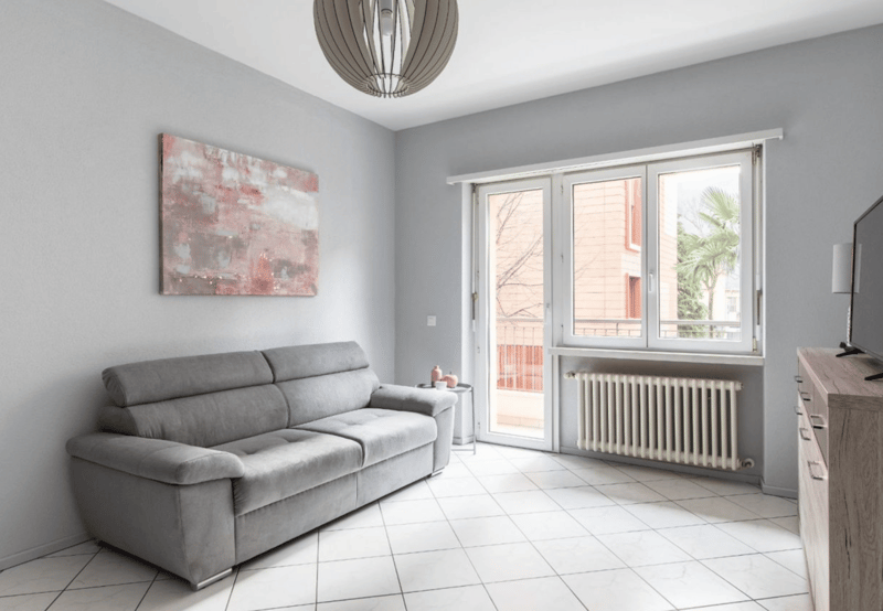 modern living room with gray couch, white tiles, white walls, large windows, balcony, radiator, painting, table, lamp
