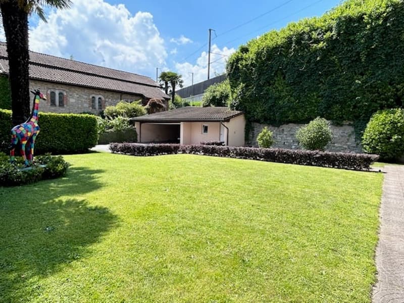 Two-story building with a tiled roof, surrounded by a well-maintained garden with a lush green lawn, hedges, and a colorful sculpture. There appears to be a covered parking area or carport on the side of the building.