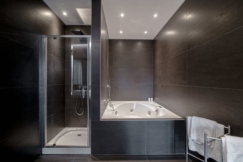 Large bathroom with dark gray tile walls, a white bathtub with jets, and a glass-enclosed shower stall. The bathroom is well-lit with recessed lighting.