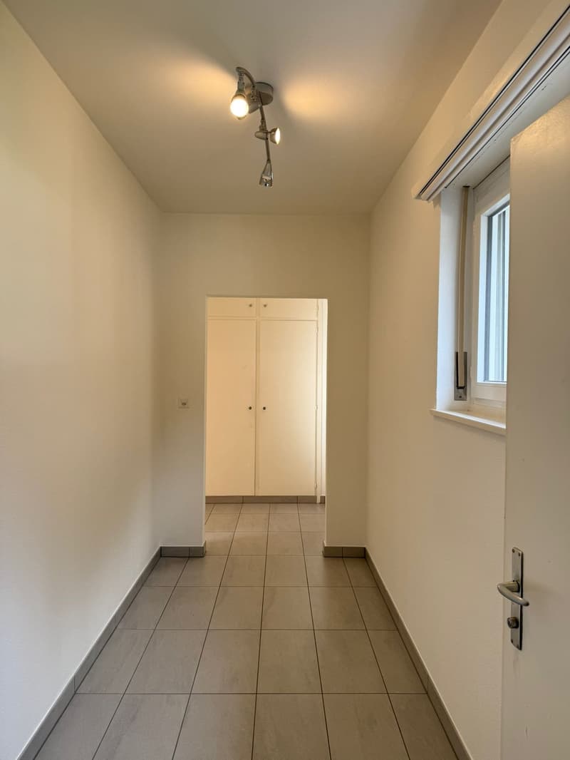 Empty hallway with white walls, tiled floor, a window, double doors, and a wall mounted lighting fixture