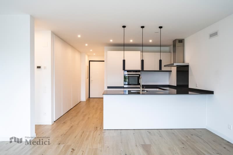 Modern kitchen with black countertops, white cabinets, wooden floor, hanging lamps, a sink with a faucet, an oven, an extractor hood, and a white central island.