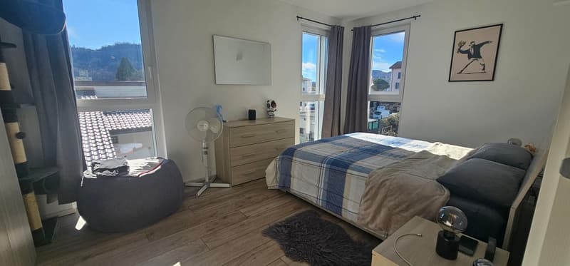 Bedroom with wooden floor, bed, nightstands, nightstand lamp, rug, chest of drawers, fan, picture frame on the wall, and window with curtain