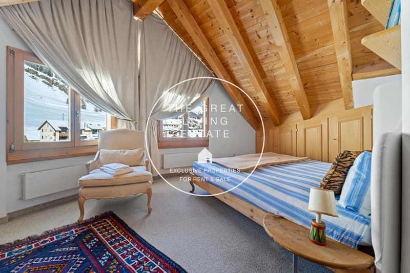 Spacious bedroom with wooden beamed ceiling, large windows overlooking a snowy landscape, a canopy bed, and a colorful patterned rug on the floor.