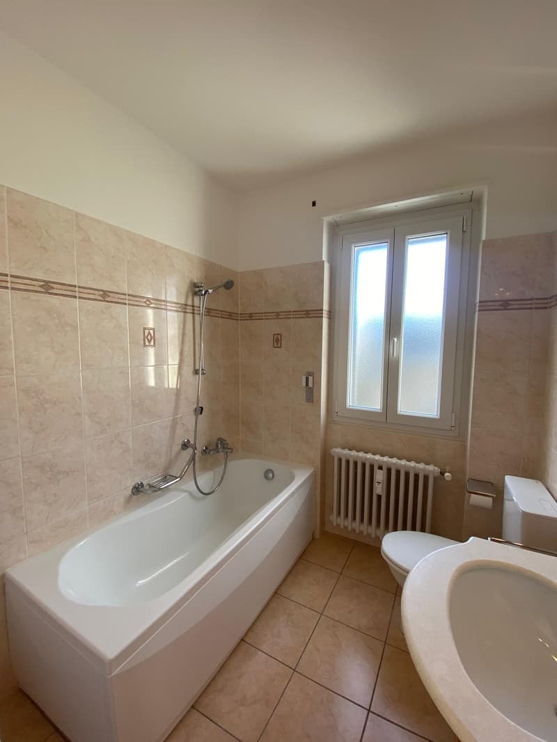Modern bathroom with a bathtub, toilet, sink, and radiator. Beige tile flooring and walls.