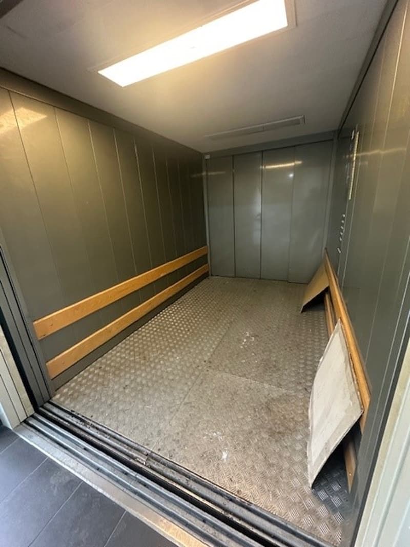 Inside an elevator, grey walls, wooden flooring, open doors, white light on the ceiling