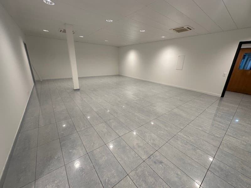 A room with white walls, ceiling, and tiled floor, and lights installed in the ceiling. There is a pillar in the middle of the room and an exit door at the right end.
