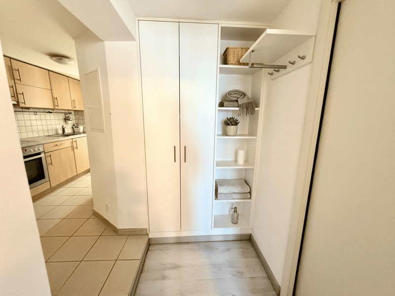 Empty room with white wardrobe, shelves, and tiled floor. Kitchen area visible in the background.