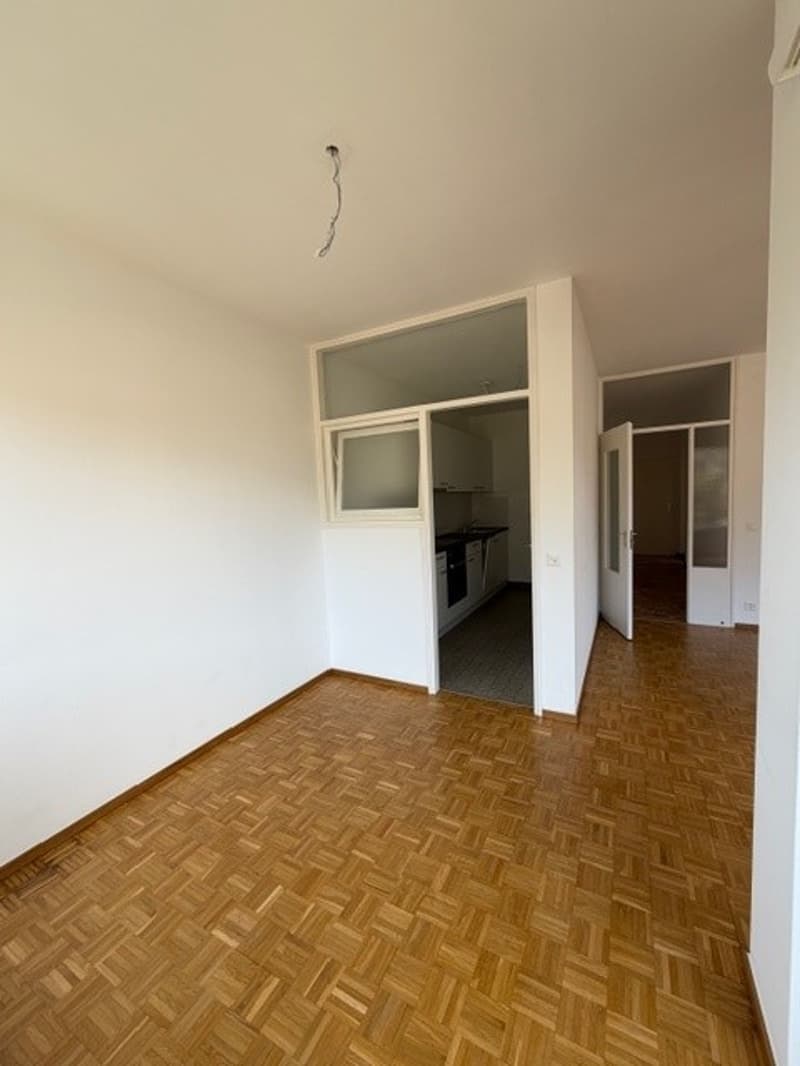 Empty room with wooden floor, white walls, open door to kitchen, windows
