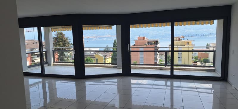 Large open-plan living space with floor-to-ceiling windows offering a panoramic view of the surrounding cityscape and lake. The room has a sleek, modern design with white tile flooring and black window frames.