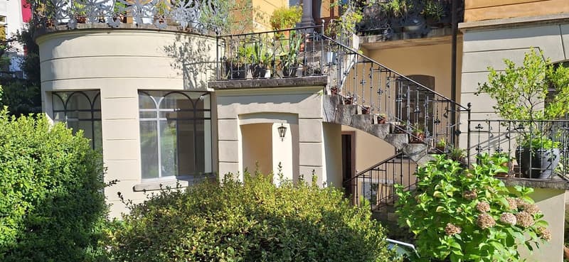2-story building with arched windows, balcony, and lush greenery surrounding the entrance