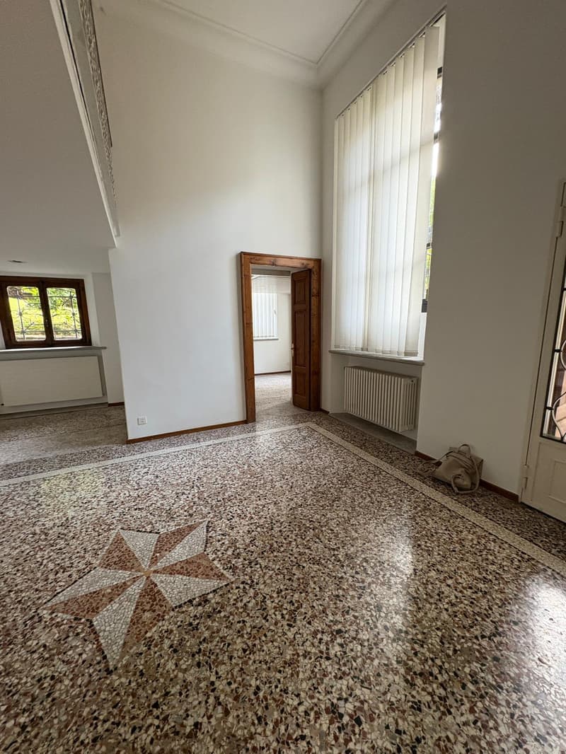 An empty hallway with mosaic flooring and white walls, with windows and doors, one of which has white curtains. There is a radiator against one wall and a star pattern in the floor.