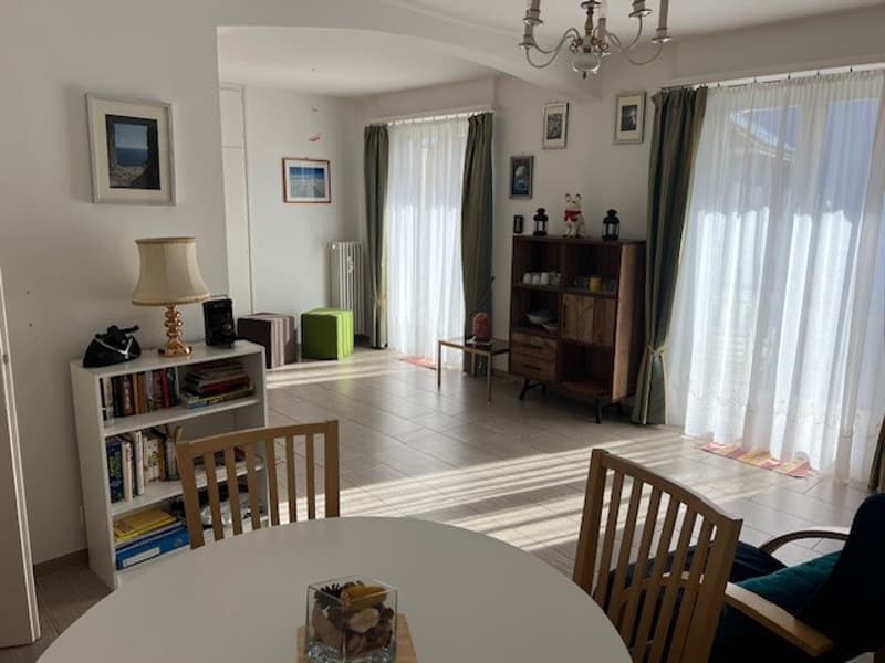 large room with white walls, white tiled floor, glass windows with white curtains, wooden dining table with glass decoration, shelves with books and items, white armchairs, wooden cabinet, lamps, chandelier on ceiling