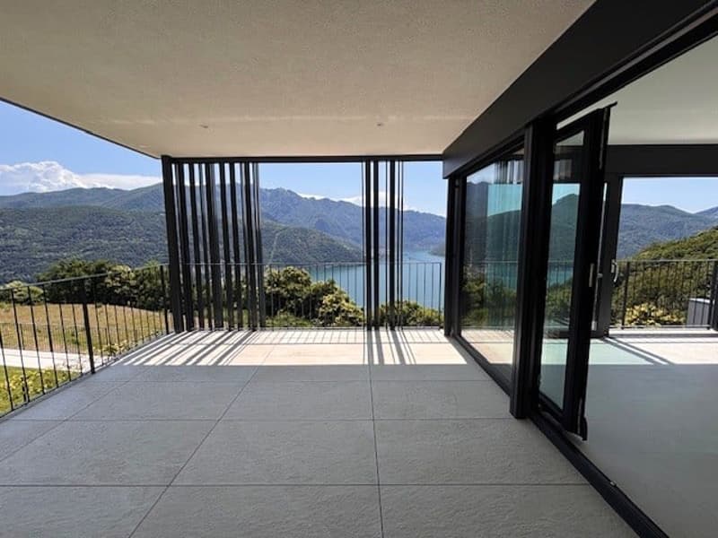Large balcony or terrace with panoramic views of a mountainous landscape and a body of water, such as a lake or ocean. The space features floor-to-ceiling glass windows and doors, providing an open and unobstructed view of the scenic surroundings.