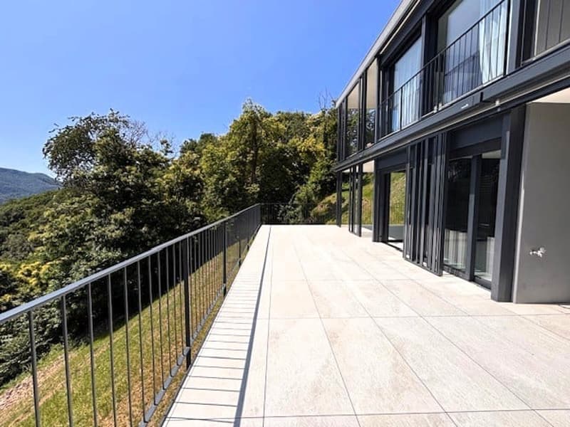 A modern building with a large balcony or terrace overlooking a lush, green landscape. The building has large windows and a sleek, contemporary design.