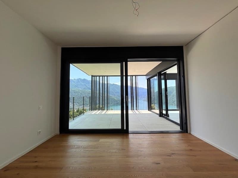 Spacious living room with large windows and sliding glass doors leading to a balcony or terrace, offering a scenic view of the surrounding mountains or landscape. The room has hardwood flooring and a minimalist, modern design.
