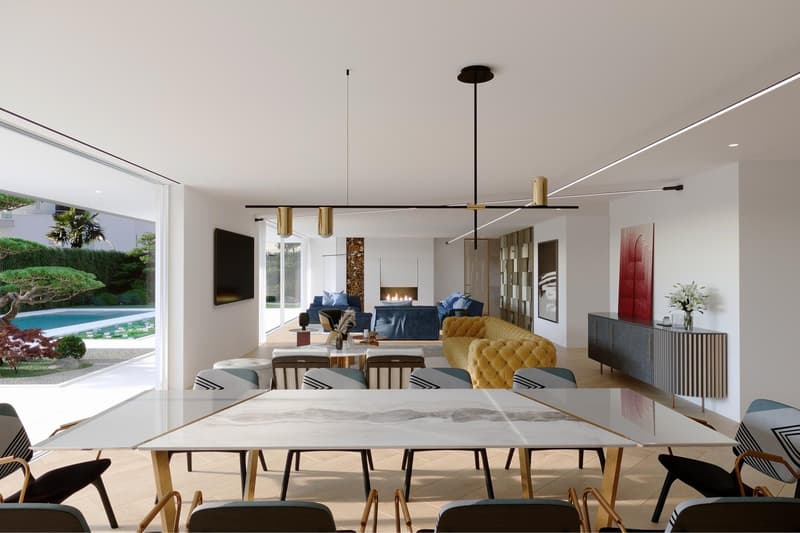 Spacious open-plan living and dining area with large windows overlooking an outdoor garden and pool. The room features a modern fireplace, a long wooden dining table with chairs, a yellow tufted sofa, and a black entertainment unit. The lighting fixtures