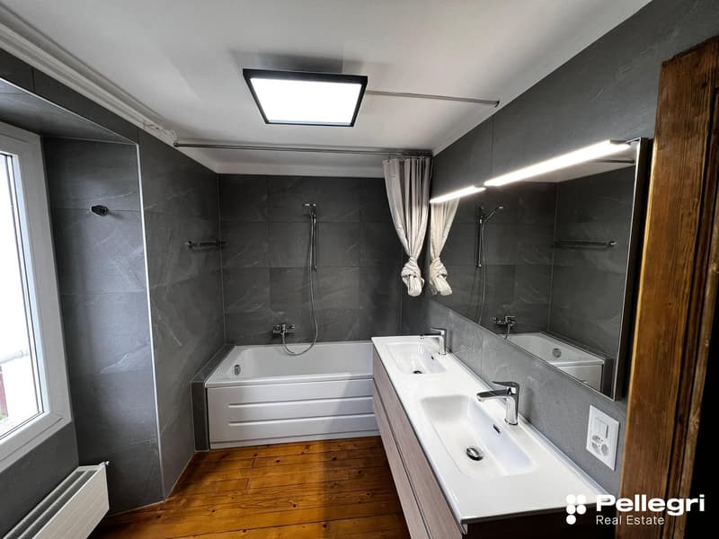Large bathroom with grey tile walls, a bathtub, a vanity with a sink, and a window. The room has modern fixtures and lighting.