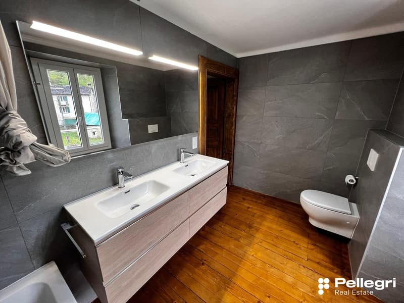 Large bathroom with grey tile walls, wooden floor, double vanity sink, and toilet