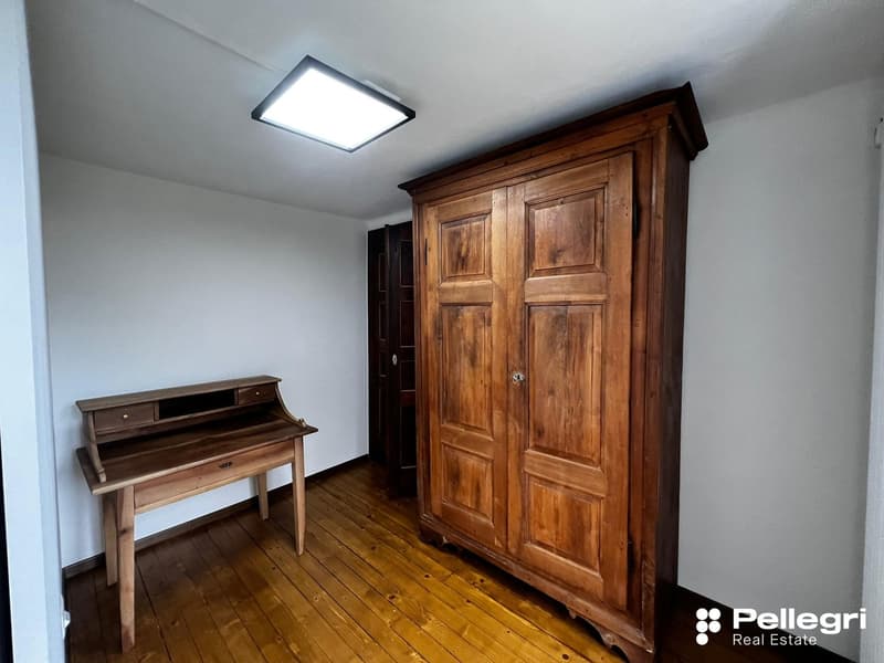 The image shows a bedroom with a large wooden wardrobe, a wooden desk, and a hardwood floor. The room has a clean, minimalist design with white walls and a bright ceiling light.