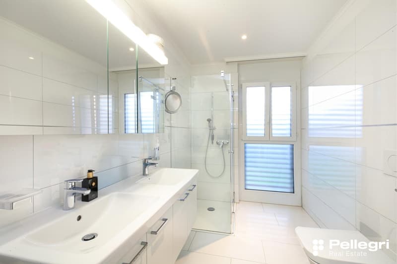 Spacious bathroom with white tiles, large vanity sink, glass shower enclosure, and large windows providing natural light