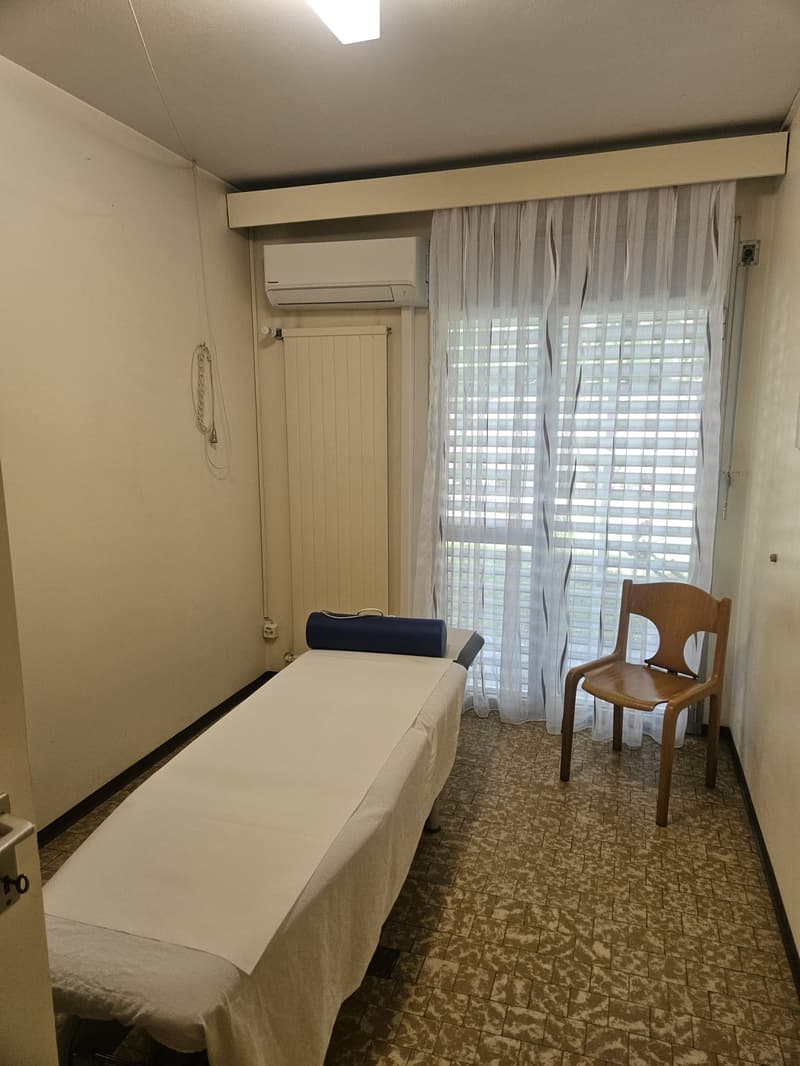 Bedroom with a massage table, a wooden chair, and a window with curtains. The room has a tiled floor and an air conditioning unit on the wall.