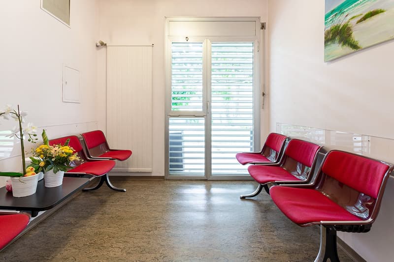 The image shows a waiting room or lobby area with red leather chairs arranged in rows. There is a large window with a view of the outdoors, and a small table with a vase of flowers. The floor appears to be a concrete or tile material.
