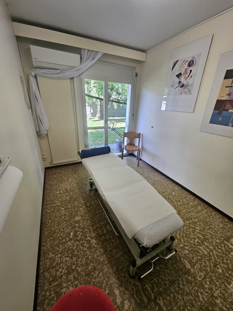 The image shows a treatment room with a massage table, a chair, and artwork on the walls. The room has large windows that provide a view of the outdoor greenery. The floor is covered with a patterned tile or linoleum material.