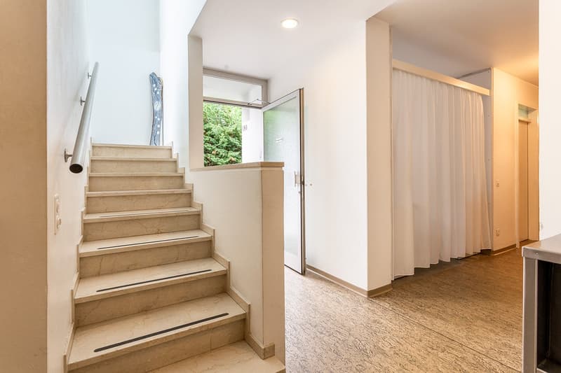 The image shows the interior of a residential property. It features a staircase with marble steps leading up to the second floor. The hallway has white walls and a curtain-covered doorway leading to another room. The floor is covered in a light-colored ca