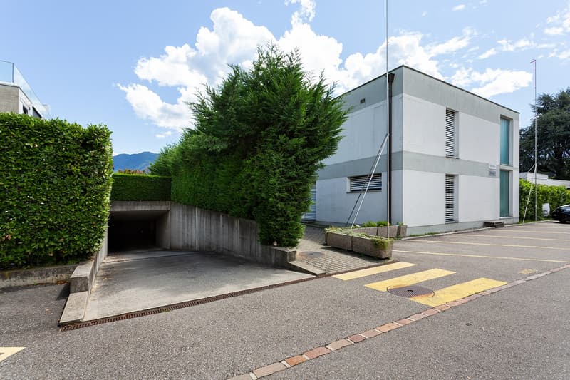 A modern, two-story commercial or industrial building with a garage entrance, surrounded by landscaping including tall hedges and trees.