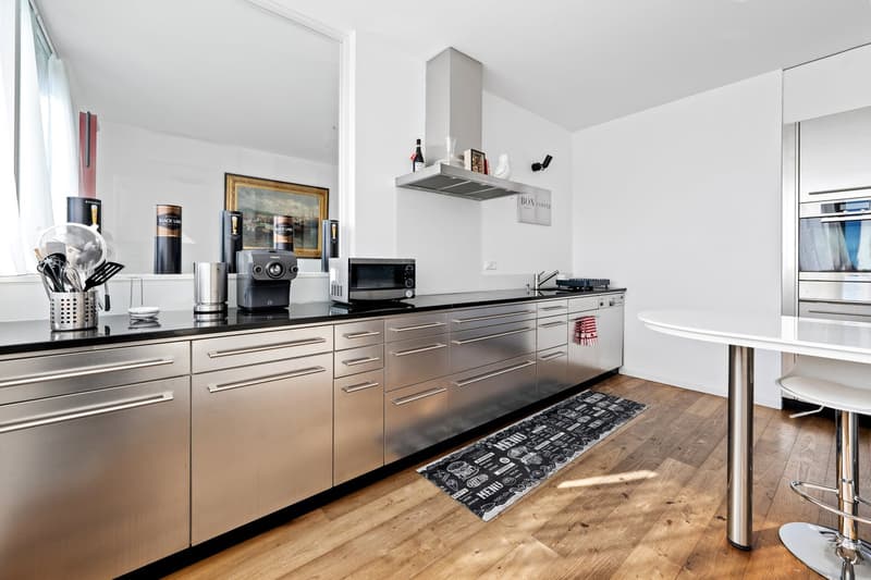 modern kitchen with stainless steel cabinets, wooden floor, kitchen appliances, large round white table, chrome bar stools, white countertop, rug