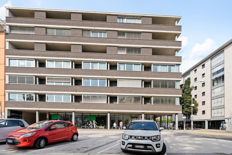 multi-story residential building with brick facade and glass windows, parking area in front, multiple vehicles parked