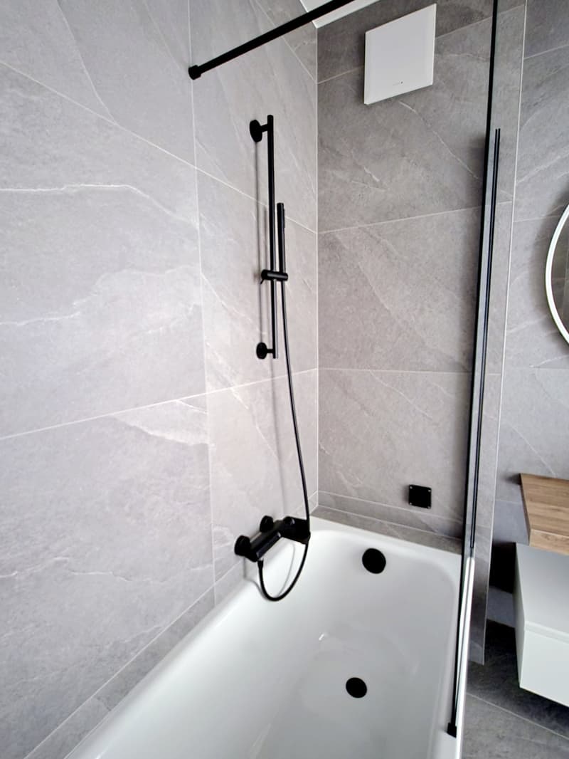Bathroom with white bathtub, glass door, gray tiles, black shower head and hose