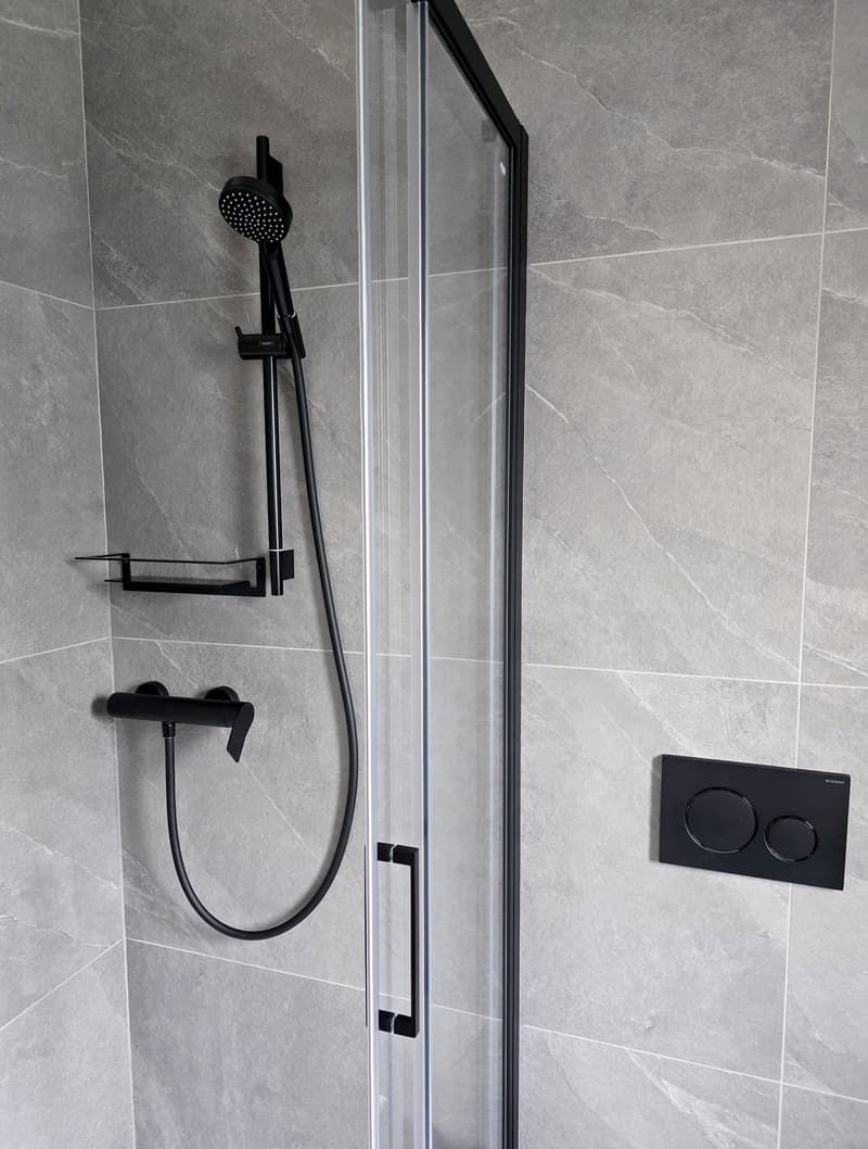 Gray tiled wall, black shower head and handle, glass door, and metal shelf.