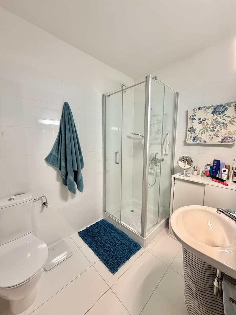 modern bathroom with white tiles, glass shower, white sink, white toilet, blue rug, towel, mirror, toiletries