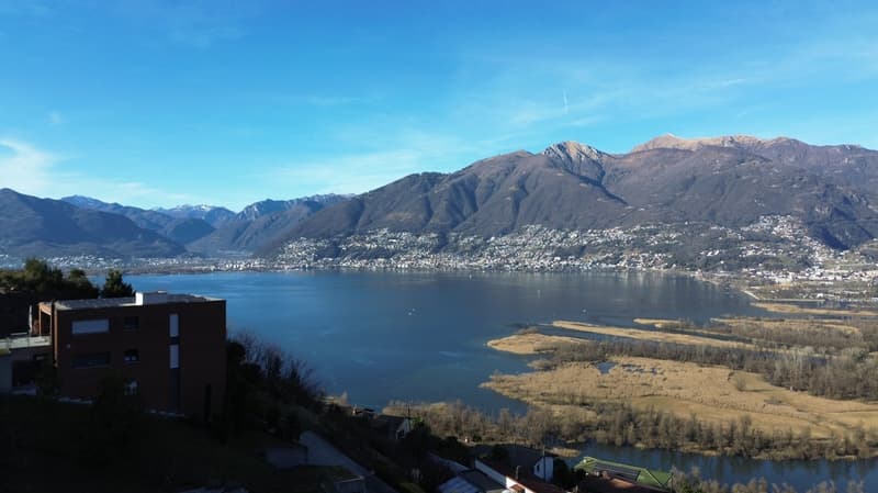 Aerial view of a mountainous region with a large lake in the foreground, a town in the distance, mountains covered with snow, a clear blue sky, and a reflection of the mountains in the water