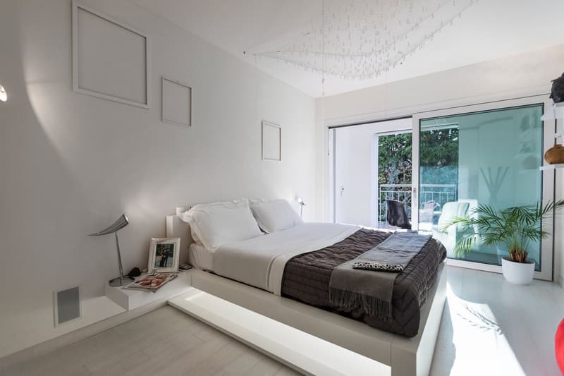 modern design, white walls, bed with white bedding, bedside table with lamp, picture frame, sliding glass door to balcony, potted plant, white flooring