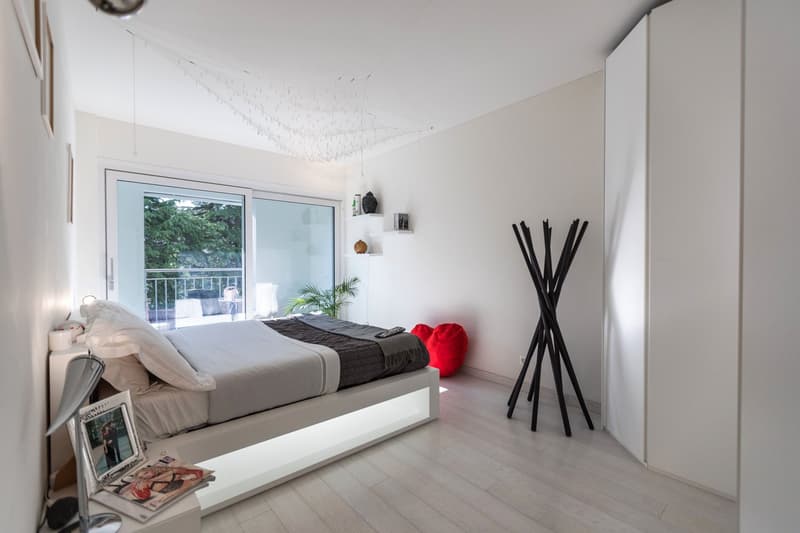 modern bedroom with white walls, wooden floor, bed with white bedding, bedside table with lamp, picture frame, magazine, and decorative items, sliding glass door leading to balcony with railing, and a plant