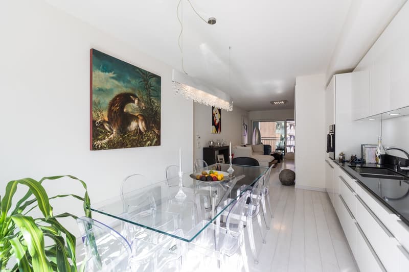 modern dining area with glass table, clear chairs, white walls, modern kitchen, white cabinets, black countertops, modern chandelier