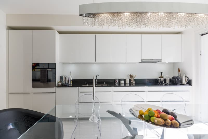 modern kitchen with white cabinets, black countertops, glass table, chandelier, clear chairs, bowl of fruits