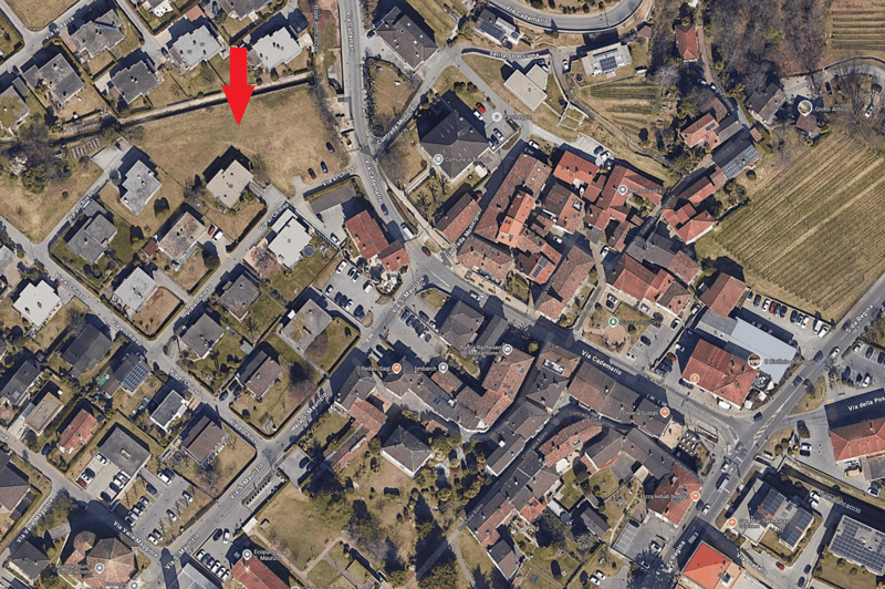 Aerial view of a neighborhood with residential houses, paved roads, and parking spaces. Notable structures include a small park, solar panels, and a roundabout.