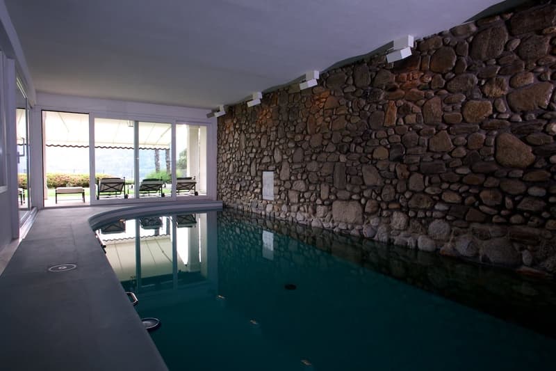 The image shows an indoor swimming pool with a stone wall on one side. The pool has a green tiled surface and is surrounded by large windows that provide a view of the outdoor area. There are chairs visible through the windows, suggesting an outdoor seati