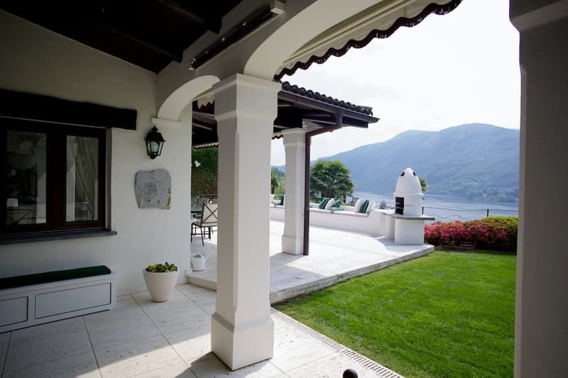 Covered outdoor patio with arched entryway, tiled floor, and views of the mountains and lake in the background. The patio has a grassy lawn, potted plants, and outdoor furniture.